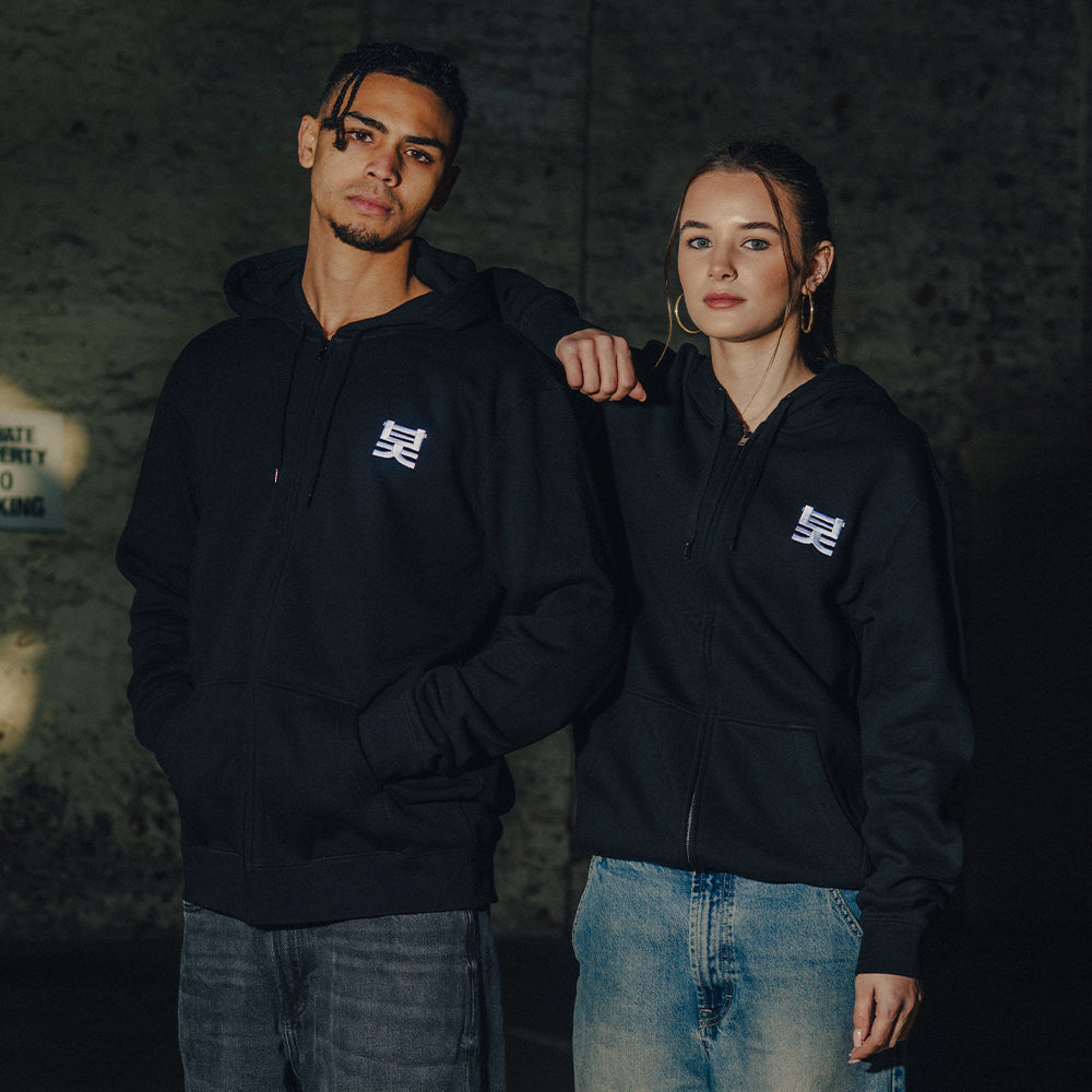 Two people wearing dark hoodies with a logo, standing close together against a dark background.